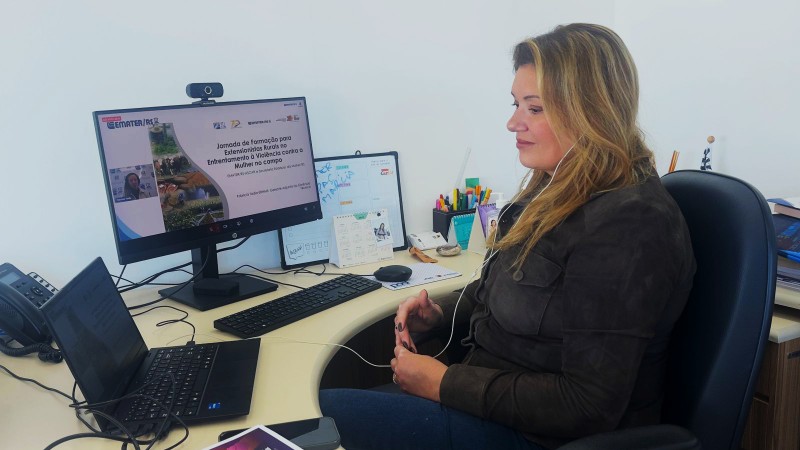 Fotografia colorida em ambiente de escrit&oacute;rio mostra uma mulher sentada diante de uma mesa, participando de uma videoconfer&ecirc;ncia. Ela usa fones de ouvido com microfone e observa atentamente o monitor do computador. Na tela, est&aacute; sendo exibida uma apresenta&ccedil;&atilde;o intitulada &ldquo;Jornada de Forma&ccedil;&atilde;o para Extensionistas Rurais no Enfrentamento &agrave; Viol&ecirc;ncia contra a Mulher no Campo&rdquo;, promovida pela Emater/RS. Sobre a mesa h&aacute; notebook, teclado, telefone, materiais de escrit&oacute;rio e objetos de uso cotidiano. A imagem registra um momento de capacita&ccedil;&atilde;o on-line voltado ao fortalecimento das a&ccedil;&otilde;es de enfrentamento &agrave; viol&ecirc;ncia contra as mulheres no meio rural.