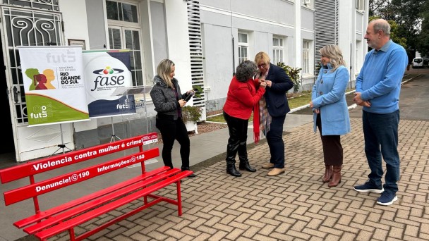 Fotografia em &aacute;rea externa, em frente a um pr&eacute;dio institucional de fachada branca. Em primeiro plano, h&aacute; um banco vermelho com as mensagens: &ldquo;Viol&ecirc;ncia contra mulher &eacute; crime!&rdquo;, &ldquo;Denuncie! 190&rdquo; e &ldquo;Informa&ccedil;&otilde;es: 180&rdquo;, s&iacute;mbolo da campanha de enfrentamento &agrave; viol&ecirc;ncia contra as mulheres. Ao lado do banco, banners da Secretaria da Mulher do Governo do Estado do Rio Grande do Sul, da Fase e da Fut5 est&atilde;o posicionados em suportes.

&Agrave; direita, cinco pessoas participam de uma cerim&ocirc;nia. No centro, uma mulher de cabelos grisalhos, usando casaco vermelho, entrega um broche &agrave; secret&aacute;ria da Mulher, Ana Costa, que veste blazer azul-marinho, len&ccedil;o estampado, cal&ccedil;a escura e sapatos bege. Outra mulher registra o momento com um celular. Um homem e uma mulher observam a cerim&ocirc;nia sorrindo. O ambiente &eacute; arborizado e transmite um contexto de homenagem e compromisso institucional com a preven&ccedil;&atilde;o e o enfrentamento da viol&ecirc;ncia contra as mulheres.